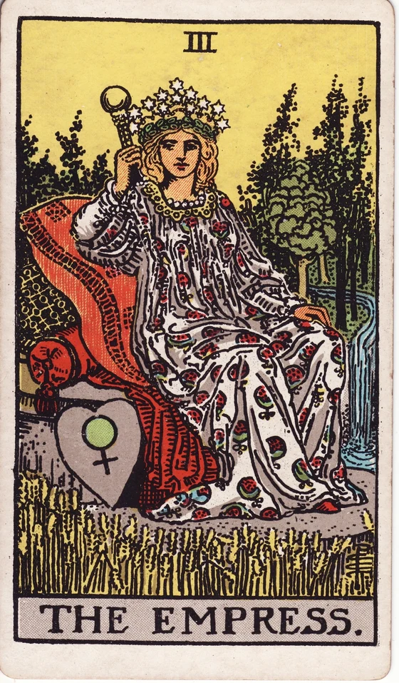 The Empress tarot card from the Rider–Waite–Smith deck: a crowned woman on cushions in a field of wheat beside a waterfall, a heart-shaped shield with the Venus sign.